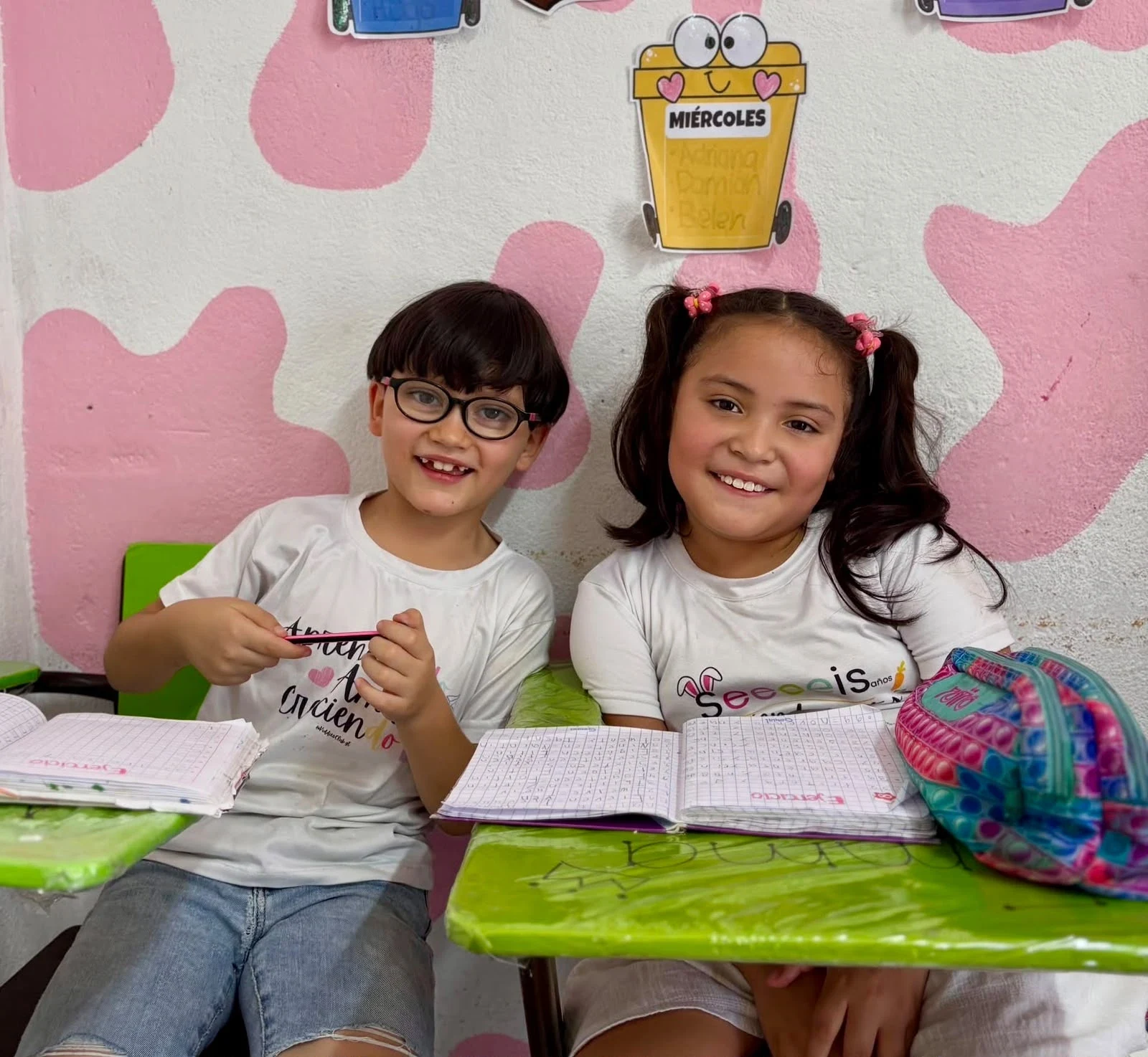 Dos compañeros de clase de Kiddies Club sonriendo en sus pupitres con sus cuadernos abiertos