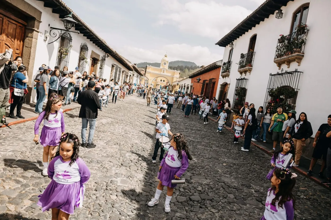 Niñas de Kiddies Club con uniforme lila desfilando por una calle empedrada, con el Arco de Santa Catalina al fondo