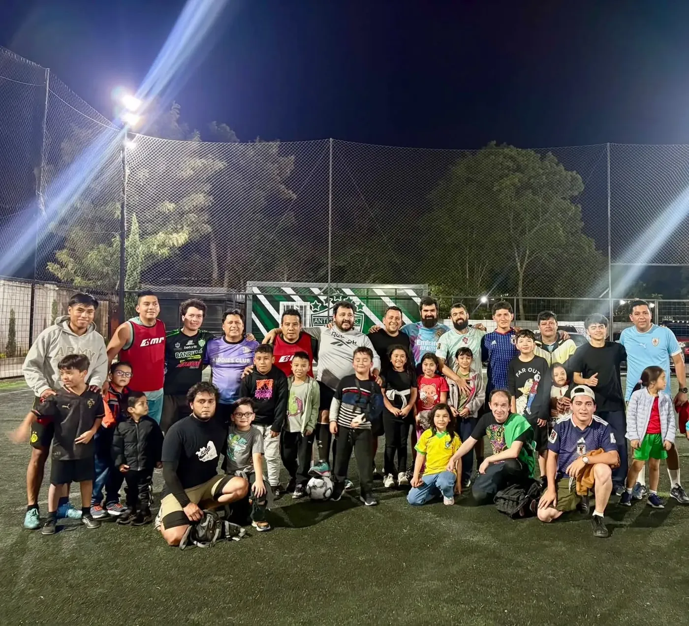 Papás y alumnos de Kiddies Club juntos en una cancha de fútbol de noche, celebrando el Día del Padre