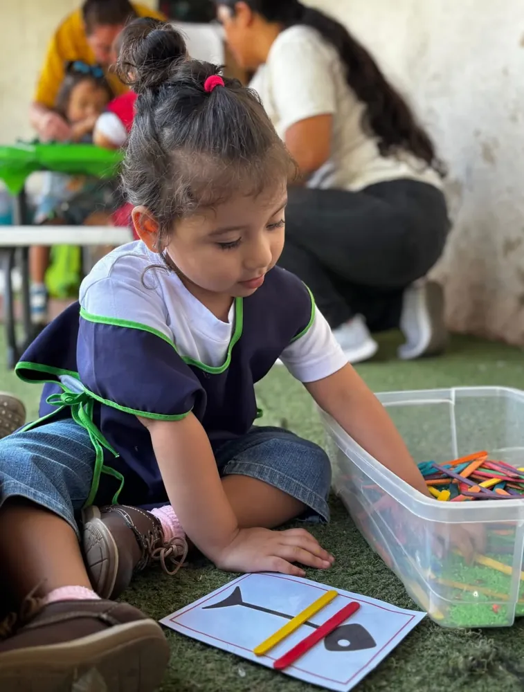 Niña pequeña de Kiddies Club en una estación sensorial, colocando palitos de colores sobre una tarjeta