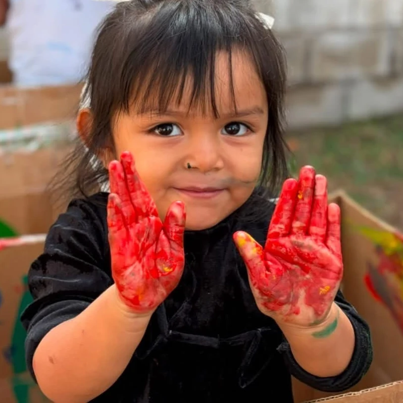 Niña pequeña de Kiddies Club sonriendo y mostrando sus manos llenas de pintura roja